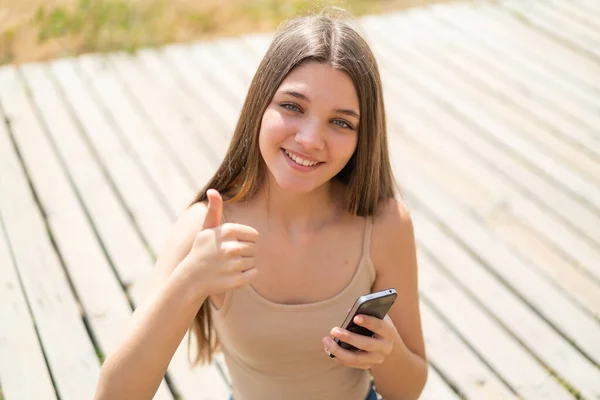 少女在户外竖起大拇指的时候用手机 — 图库照片