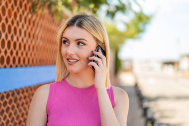 Açık havada genç, güzel sarışın bir kadın cep telefonuyla biriyle sohbet ediyor.