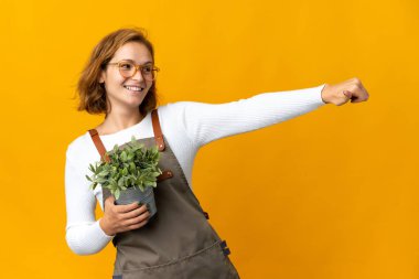 Sarı arka planda izole edilmiş bir bitkiyi tutan genç Gürcü kadın baş parmak işareti yapıyor.