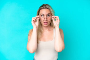 Mavi arka planda gözlüklü ve şaşkın bakışlı genç beyaz kadın.