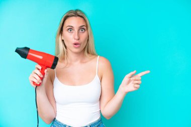 Mavi arka planda saç kurutma makinesi tutan beyaz bir kadın şaşırmış ve işaret ediyor.