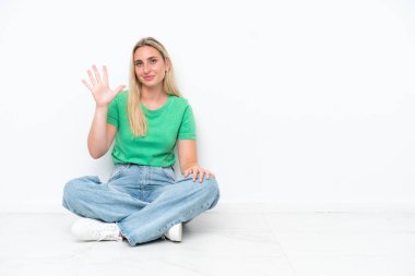 Beyaz arka planda yerde oturan beyaz tenli genç kadın parmaklarıyla beşe kadar sayıyor.