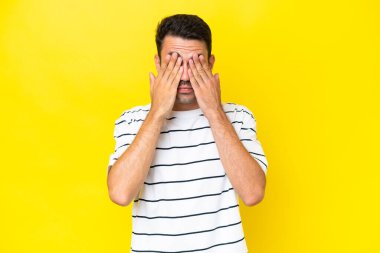 Elleri ile gözleri kaplayan izole sarı arka plan üzerinde genç yakışıklı adam