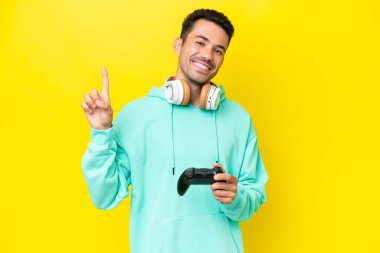 Genç yakışıklı bir adam, izole edilmiş duvarın üzerinde bilgisayar oyunu kontrolörüyle oynuyor ve en iyinin işareti olarak parmağını kaldırıyor.