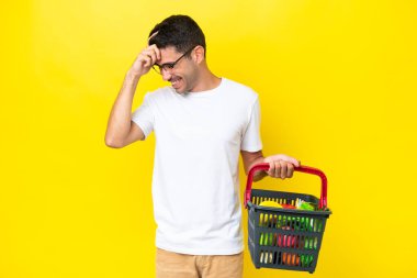 Genç yakışıklı bir adam elinde yiyecek dolu bir sepetle izole edilmiş sarı arka planda gülüyor.