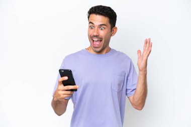 İzole edilmiş beyaz arka plan üzerinde genç yakışıklı bir adam kameraya bakarken şaşırmış bir ifadeyle cep telefonuna bakıyor.