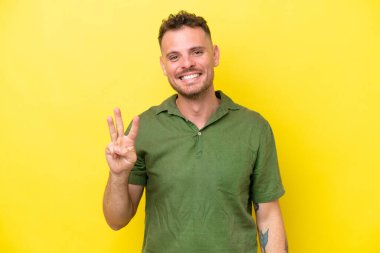 Beyaz tenli yakışıklı adam sarı arka planda izole edilmiş mutlu ve parmaklarıyla üçe kadar sayıyor.