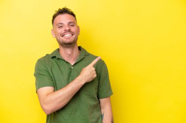 Sarı arka planda izole edilmiş beyaz, yakışıklı genç bir adam bir ürün sunmak için kenarı işaret ediyor.