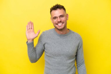 Beyaz tenli yakışıklı adam sarı arka planda izole edilmiş mutlu bir ifadeyle selamlıyor.