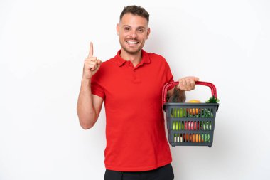 Beyaz arka planda izole edilmiş yiyecek dolu bir sepet taşıyan genç beyaz bir adam en iyinin işaretini gösteriyor ve parmağını kaldırıyor.
