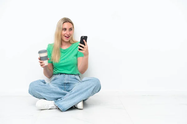 白い背景にコーヒーを持って隔離された床の上に座っている若い白人女性を取り上げ モバイル — ストック写真