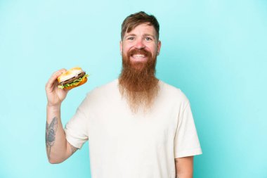 Kızıl saçlı, uzun sakallı, elinde bir burger tutan, mavi arka planda izole edilmiş, gülümseyen bir adam.