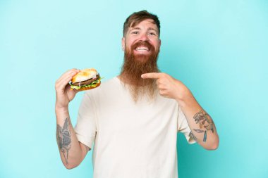 Kızıl saçlı, uzun sakallı bir adam elinde bir burger tutuyor mavi arka planda izole edilmiş ve onu işaret ediyor.