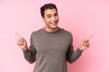 Pembe arka planda izole edilmiş genç beyaz adam parmak uçlarını işaret ediyor ve mutlu.