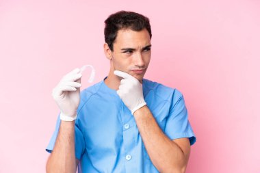 Dentist caucasian man holding invisible braces isolated on pink background having doubts and thinking