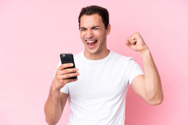 Young caucasian man isolated on pink background using mobile phone and doing victory gesture