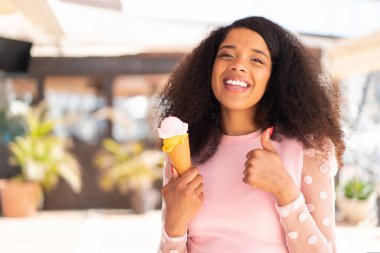 Açık havada kornet dondurmalı genç Afrikalı Amerikalı bir kadın. Başparmakları havada. Çünkü iyi bir şey oldu.