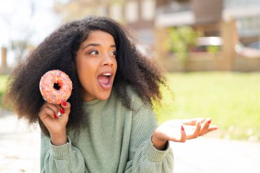 Genç Afrikalı Amerikalı bir kadın açık havada yüzünde sürpriz bir ifadeyle donut tutuyor.
