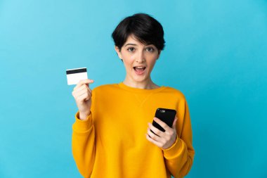 Kısa saçlı bir kadın mavi arka planda izole edilmiş cep telefonuyla alışveriş yapıyor ve elinde şaşırmış bir ifadeyle bir kredi kartı tutuyor.