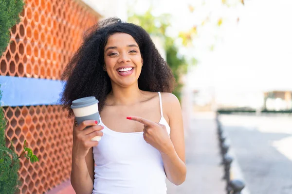 Genç Afro-Amerikalı bir kadın açık havada kahve tutuyor ve onu doğrultuyor.