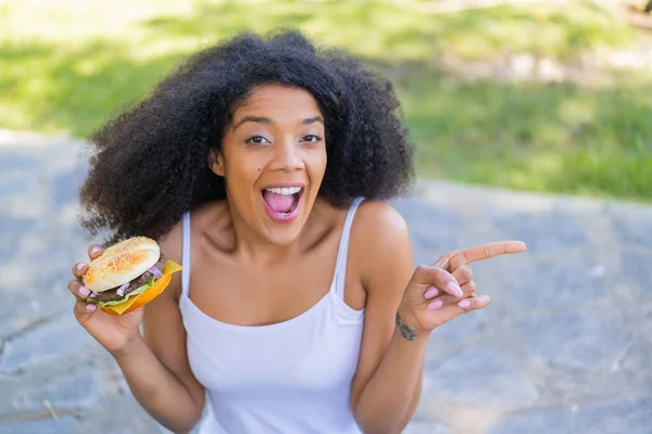 Młoda Afroamerykanka Trzyma Hamburgera Świeżym Powietrzu Zaskoczony Wskazując Palcem Bok — Zdjęcie stockowe