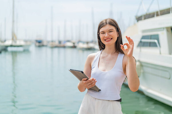 Young pretty Ukrainian woman holding a tablet at outdoors showing ok sign with fingers