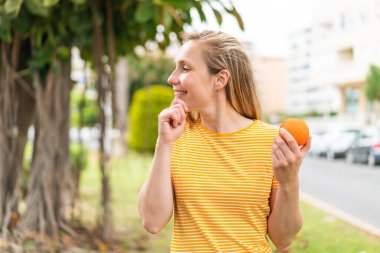 Açık havada elinde portakal tutan genç sarışın kadın bir fikir üretiyor ve ona bakıyor.