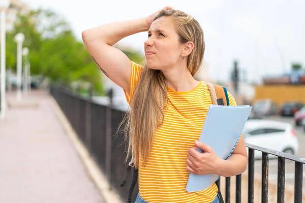 Young blonde student woman at outdoors having doubts and with confuse face expression