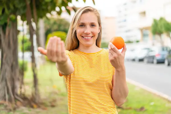 Young blonde woman holding an orange at outdoors inviting to come with hand. Happy that you came