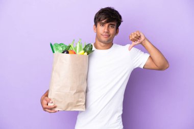 Elinde bir alışveriş çantası tutan genç adam mor arka planda izole edilmiş, olumsuz bir ifadeyle baş parmağını gösteriyor.