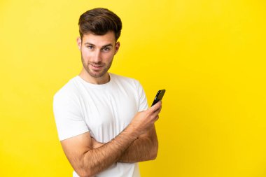Sarı arka planda izole edilmiş genç beyaz bir adam cep telefonu tutuyor ve kollarını kavuşturuyor.