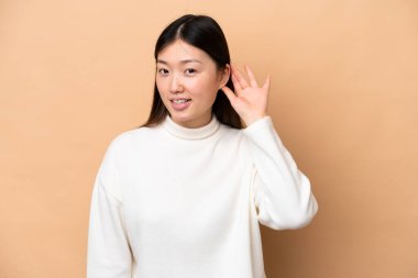 Young Chinese woman isolated on beige background listening to something by putting hand on the ear
