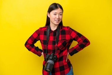 Sarı arka planda izole edilmiş genç fotoğrafçı kadın kolları kalçasında ve gülümsüyor.