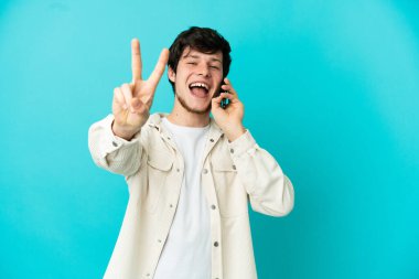 Mavi arka planda izole edilmiş cep telefonu kullanan genç Rus adam gülümsüyor ve zafer işareti gösteriyor.