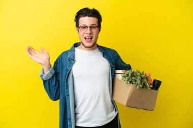 Adam sarı arka planda izole edilmiş ve şok olmuş yüz ifadesi olan şeylerle dolu bir kutuyu kaldırırken hamle yapıyor.