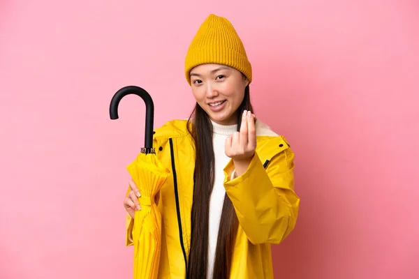 Young Chinese woman with rainproof coat and umbrella isolated on pink background making money gesture