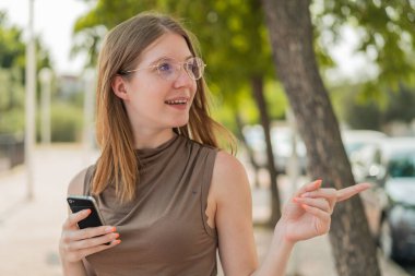 Gözlüklü Fransız kız, açık havada cep telefonu kullanıyor. Ürünü sunmak için yan tarafı işaret ediyor.