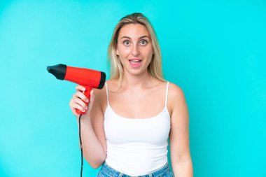 Mavi arka planda saç kurutma makinesi tutan beyaz bir kadın. Şaşırmış ve şok olmuş bir yüz ifadesi var.