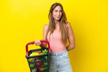 Young caucasian woman holding a shopping basket full of food isolated on yellow background and looking up