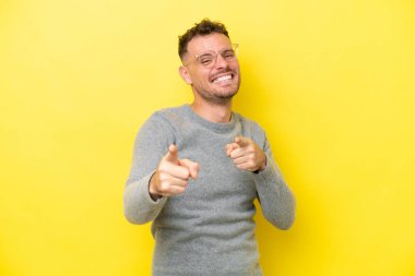 Sarı arka planda izole edilmiş beyaz, yakışıklı genç adam şaşırmış ve ön tarafı işaret ediyor.