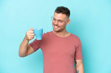 Kafkasyalı genç bir adam elinde bir fincan kahve tutarken mavi arka planda mutlu bir ifadeyle izole edilmiş.