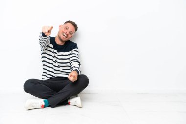 Genç bir adam beyaz arka planda izole bir şekilde yerde oturuyor. Gülümserken seni işaret ediyor.