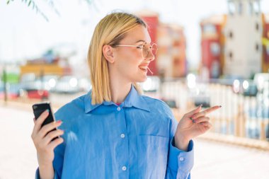 Açık havada cep telefonu kullanan genç, güzel sarışın bir kadın bir ürün sunmak için yan tarafı işaret ediyor.