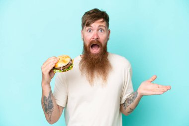 Kızıl saçlı, uzun sakallı bir adam elinde hamburgerle mavi arka plandan izole edilmiş, şok olmuş bir yüz ifadesi var.