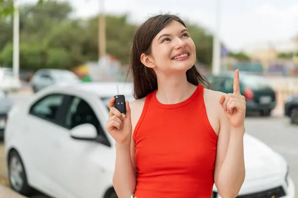若い美しいウクライナの女性は 指を上げながら解決策を実現しようと屋外で車のキーを保持しています — ストック写真