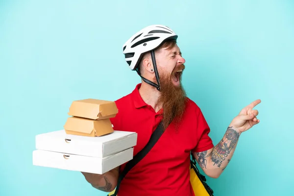 Delivery man holding pizzas and burgers isolated on blue background pointing finger to the side and presenting a product
