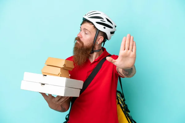 Delivery man holding pizzas and burgers isolated on blue background making stop gesture and disappointed
