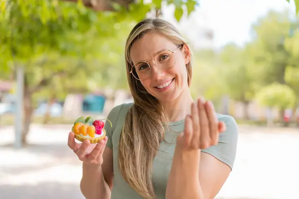 Young blonde woman holding a tartlet at outdoors inviting to come with hand. Happy that you came