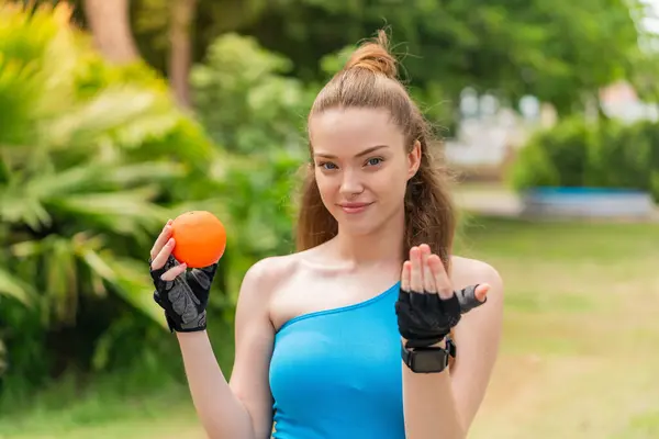 Young pretty sport girl holding an orange at outdoors inviting to come with hand. Happy that you came