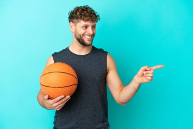 Basketbol oynayan yakışıklı genç adam mavi arka planda izole edilmiş bir şekilde parmağını yana uzattı ve bir ürün sundu.